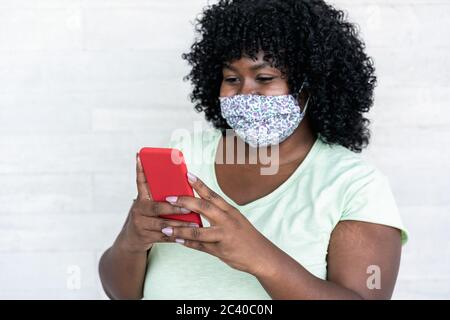 Happy black girl con smartphone outdoor - giovane donna africana curvy divertirsi guardando i video sul telefono cellulare mentre si indossa la maschera protettiva per il viso - Foto Stock