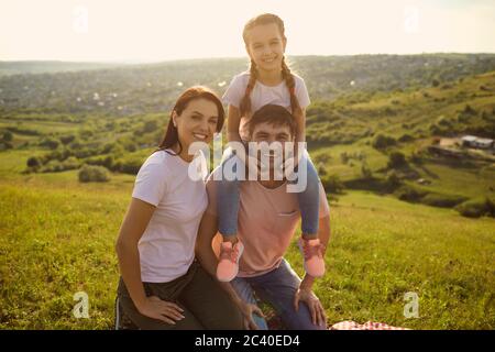 La famiglia è felice di trascorrere il tempo insieme nella natura al tramonto. Ritratto di carina ragazza e dei suoi genitori in vacanza estiva Foto Stock