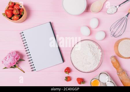 Composizione piatta composizione di posa taccuino vuoto accanto agli ingredienti per cuocere la torta di fragole, utensili e fiori freschi su tavola rosa. Foto Stock