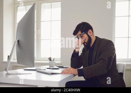 Esausto lavoratore stanco sul posto di lavoro ha coperto il suo volto con le mani in ufficio Foto Stock