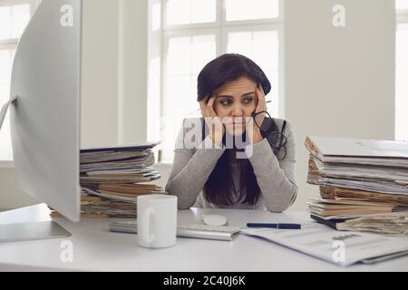 Donna bruna ragioniere in occhiali con una montagna di documenti sul tavolo stanco depresso emaciato depressivo deluso al tavolo nel Foto Stock