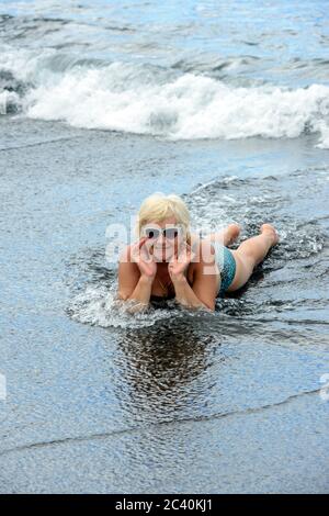 Ritratto di donna anziana che si sta adagiando sul bordo del surf su sabbia vulcanica nera bagnata della spiaggia Arena a Puerto de Santiago, Tenerife, Canary Islan Foto Stock