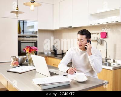 giovane uomo asiatico di affari seduto cucina contatore che lavora a casa guardando computer portatile che parla su telefono cellulare Foto Stock