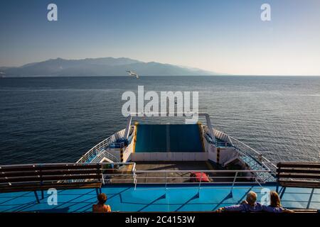 Una coppia e una donna sola seduti sul ponte di un traghetto sul mare aperto in una giornata estiva soleggiata, un gabbiano che vola sopra, montagne davanti, Thassos Isl Foto Stock