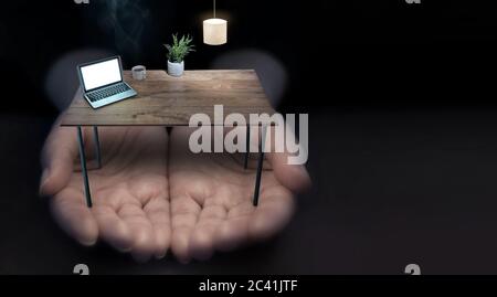Mani allungate che tengono una scrivania da ufficio in miniatura con computer portatile, lampada e caffè caldo Foto Stock