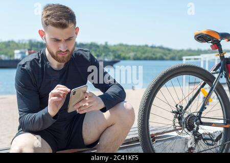 Ritratto di un atleta di ciclista che riposa dopo un giro in bicicletta usando uno smartphone in mano. All'aperto. Sport primaverili. Il ragazzo ha venticinque anni Foto Stock