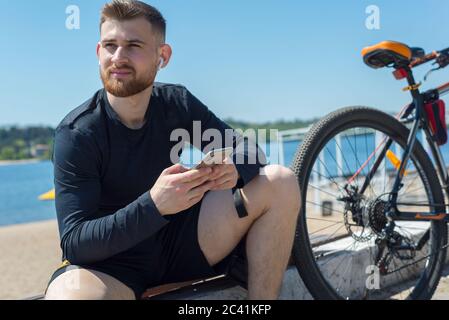 Ritratto di un atleta di ciclista che riposa dopo un giro in bicicletta usando uno smartphone in mano. All'aperto. Sport primaverili. Il ragazzo ha venticinque anni Foto Stock