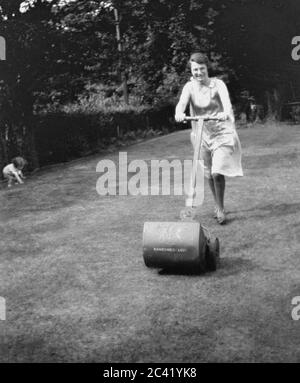 Anni '30, storica, una signora che taglia l'erba in un giardino posteriore con un'erba che si fa mormorare dell'epoca, con il suo bambino che gioca all'angolo, Inghilterra, Regno Unito. Sta usando un tosaerba a spinta manuale realizzato da Ransomes che ha fatto il primo tosaerba del mondo nel 1832, sotto licenza da un'invenzione due anni ealier nel 1830 da Edward Beard che accudise un ingegnere da Stroud, Gloucestershire, Inghilterra. Foto Stock