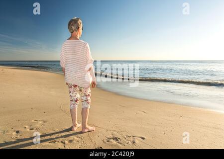 Donna anziana in piedi sulla spiaggia e guardando verso l'oceano Foto Stock