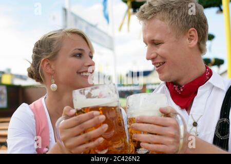 Giovane donna e giovane uomo in abiti tradizionali, bicchieri con stuoie di birra - Oktoberfest - Monaco - birreria all'aperto - festival folk Foto Stock