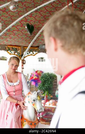 Giovane bionda a dirndl si siede su un cavallo su una giostra e flirts con un giovane uomo - Oktoberfest - Muenchen - Wiesn Foto Stock