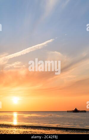 Il tramonto in un cielo parzialmente nuvoloso sul mare con i resti della testa del molo di Herne Bay sizati all'orizzonte. Il sole affonda in un cielo arancione e blu. Foto Stock