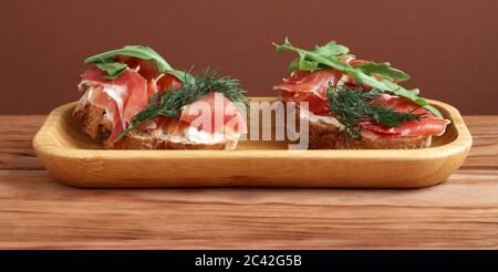 Tostature di grano saraceno con formaggio di capra, prosciutto e rucola adagiate su un piatto di bambù su un tavolo di legno. Foto Stock