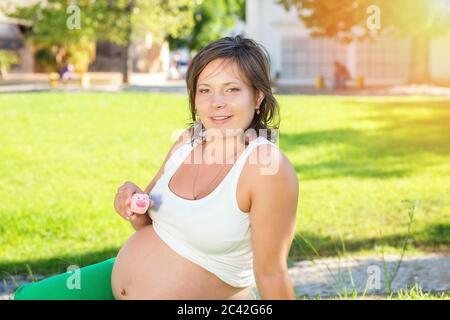 Una mamma incinta per tenere una piccola banca piggy seduta su prato verde erba nel parco all'aperto. Piccolo budget per il concetto di periodo di gravidanza. Foto Stock