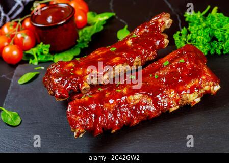 Succulente costolette di maiale al barbecue al forno con pomodoro, salsa e prezzemolo su tavola nera. Foto Stock