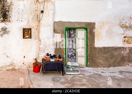 Diverse paia di scarpe sono in vendita su un tavolo davanti a una porta decorata in modo elaborato a Bhalil vicino a Fès, in Marocco, un passeggino per bambini e un secchio rosso sono accanto. Foto Stock