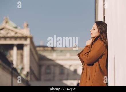 Giovane donna in cappotto marrone toffee appoggiata al muro Foto Stock