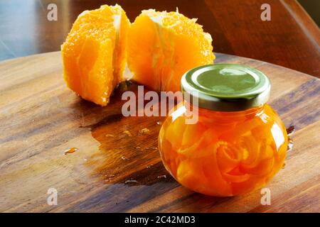 Un piccolo vasetto di marmellata fatto da bucce d'arancia è seduto su un tagliere di legno. Due metà di un arancio pelato sono sul retro. Foto Stock