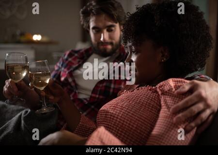 Uomo bearded abbracciando la ragazza nera e proponendo toast mentre riposava sul divano durante la cena romantica in serata a casa Foto Stock