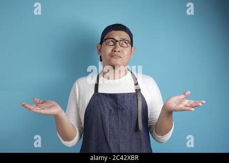 Ritratto di chef asiatico maschile o cameriere che indossa grembiule mostra le scelte tra i suoi palmi di mano vuoto, copia spazio su sfondo blu, scelta decisione Foto Stock