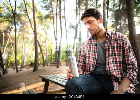 Modello uomo detiene un alluminio riutilizzabili bottiglia per acqua seduti in un parco con alberi secolari. Foto Stock