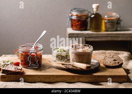 Vasetto di vetro di pate di fegato di pollo fatto in casa con pane di segale a fette, pomodori secchi e insalata di germogli verdi su tavolo da cucina in legno. Colazione a domicilio o a. Foto Stock