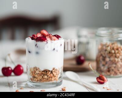 Kefir probiotico fermentato o yogurt in vetro con granola in fondo servito fresco metà ciliegia dolce su tavola rustica di legno bianco. Foto Stock