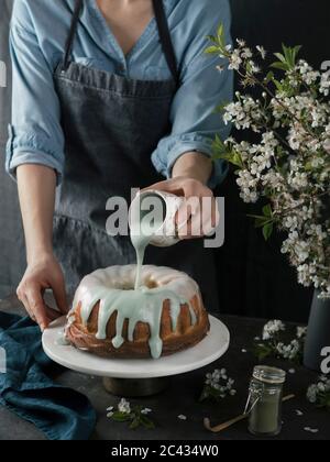 Giovane donna in camicia blu e grembiule grigio che versa una vernice verde matcha su torta a fascio su sfondo scuro. Torta di zia su supporto di torta di marmo e tappetino verde di tae Foto Stock