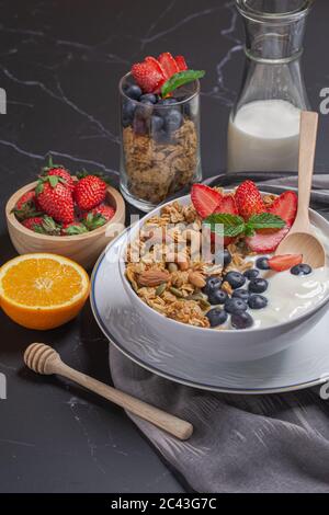 Ciotola di yogurt granola fatto in casa con frutti di bosco freschi, fragola, arancia e latte sul tavolo nero per la colazione Foto Stock