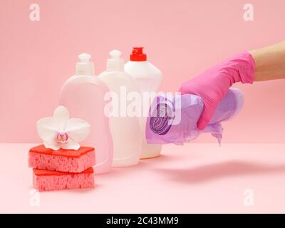 Bottiglie di plastica di liquido per lavastoviglie, spugne rosse, un fiore bianco di orchidee e una mano in un guanto di nitrile che tiene i sacchetti di spazzatura sullo sfondo rosa. W Foto Stock