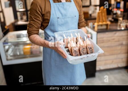 Venditore che tiene un vassoio con gelato al cioccolato su un bastone di fronte al banco del negozio, primo piano su gelato su uno sfondo blu grembiule Foto Stock