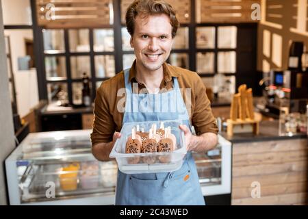 Ritratto di un allegro venditore in grembiule che tiene un vassoio pieno di gelato al cioccolato su un bastone mentre si alza in piedi la pasticceria Foto Stock