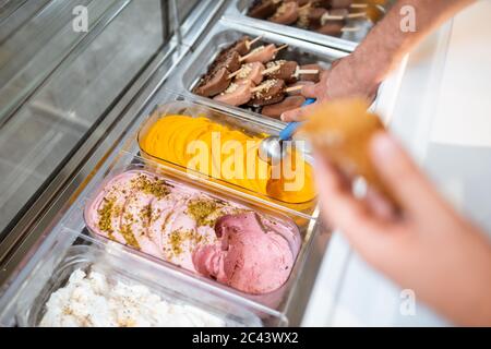 Venditore che raccoglie il gelato con un cucchiaio dal frigorifero nel negozio. Vista dall'alto su un vassoio pieno di gelato con sapori diversi Foto Stock