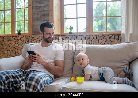 Bambino simpatico che gioca sul divano, suo padre che tiene uno smartphone e sorridente Foto Stock