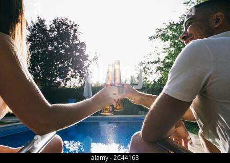 Una giovane coppia che brinda una bottiglia di birra mentre si siede a bordo piscina Foto Stock