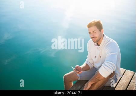 Ritratto di giovane sorridente con smartphone seduto sul molo al lago Foto Stock