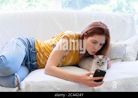 Giovane donna che si steso sul divano a casa con il suo gatto con smartphone Foto Stock