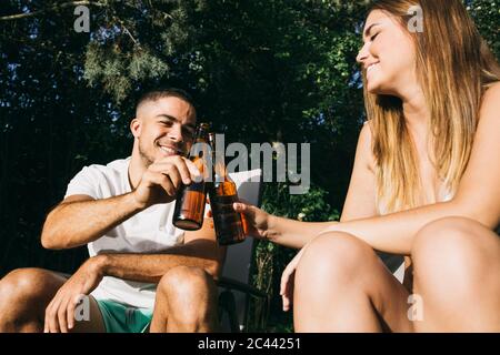 Sorridi una giovane coppia che brinda le bottiglie di birra mentre si siede al resort turistico Foto Stock