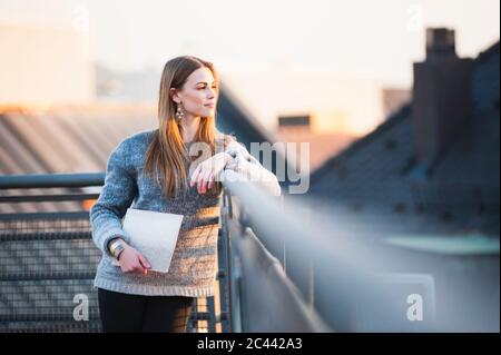 Donna giovane e premurosa che tiene il computer portatile mentre si trova sulla terrazza della città Foto Stock