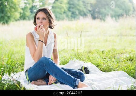 Bella giovane donna che mangia mela mentre si siede su coperta da picnic al parco Foto Stock