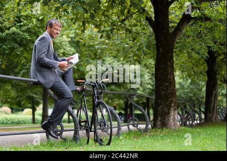 Uomo d'affari che legge giornale mentre siedono sulla ringhiera nel parco pubblico Foto Stock