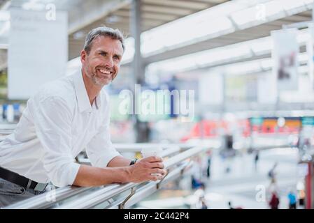 Uomo d'affari sorridente che tiene il caffè guardando via mentre si sta in piedi dalla ringhiera Foto Stock