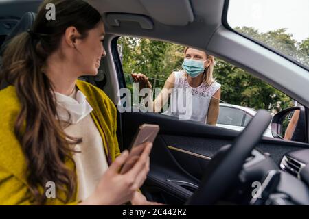 Donna che indossa una maschera protettiva che parla attraverso la finestra dell'auto con una donna senza maschera Foto Stock