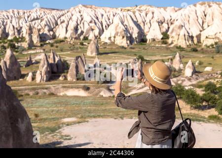 Vista laterale della giovane donna fotografare il paesaggio roccioso con smart phone al villaggio di Goreme, Cappadocia, Turchia Foto Stock