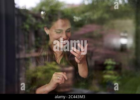 Donna mid adulta sorridente che usa il telefono cellulare a casa visto attraverso la finestra Foto Stock