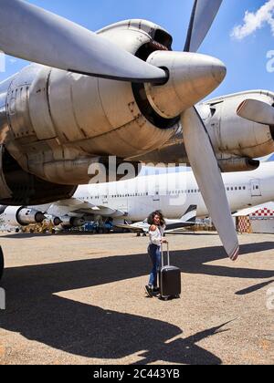 Tutta la lunghezza della giovane donna che cammina con i bagagli sotto l'aereo ad elica sulla pista dell'aeroporto Foto Stock