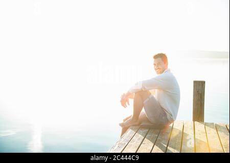 Ritratto di giovane sorridente seduto sul molo al lago dall'alba Foto Stock