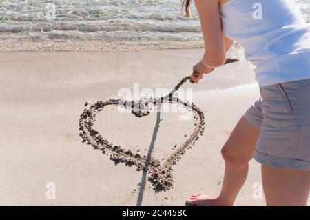 Vista raccolto di donna sulla spiaggia grattando cuore in sabbia bagnata con bastone di legno, Sardegna, Italia Foto Stock