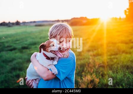 Allegro ragazzo che trasportano il cane mentre si sta in piedi sul campo durante la giornata di sole, Polonia Foto Stock