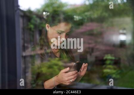 Donna mid adulta sorridente che usa lo smartphone a casa visto attraverso la finestra Foto Stock